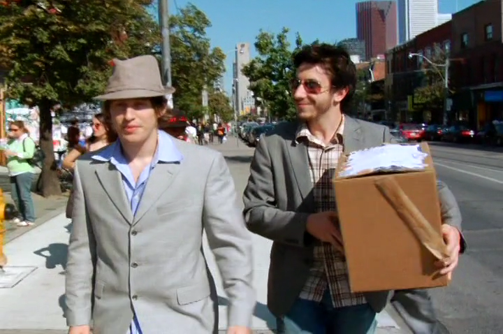 Matt and Jay walking around town. Matt is looking foward and Jay is smilling, looking at him while holding a large box, their press packgage.